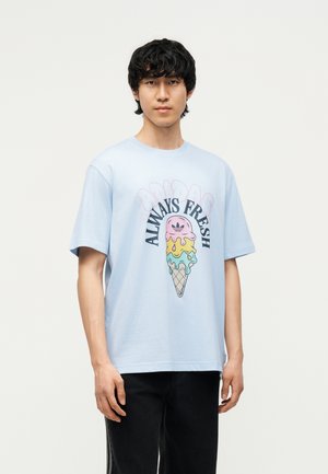 Fiatal fekete hajú férfi világoskék pólóban, amelyen színes, olvadó fagylalttölcsér és az „Always Fresh” felirat látható.