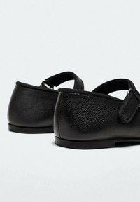 Chaussures Mary Jane en cuir noir texturé avec talons arrondis et brides ajustables sur fond blanc.