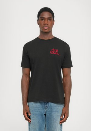 Black cotton t-shirt with crew neck, short sleeves, and red embroidered text on the left chest. Worn with light denim jeans.