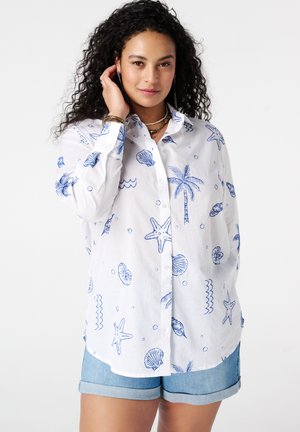 Kroeshaar vrouw draagt een wit overhemd met blauwe zee-thema print en spijkershorts, poseert met hand bij gezicht.