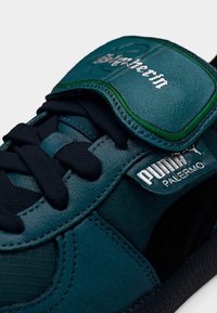 Teal leather sneaker with a textured surface, black laces, and a padded tongue featuring silver branding. Includes a green accent.