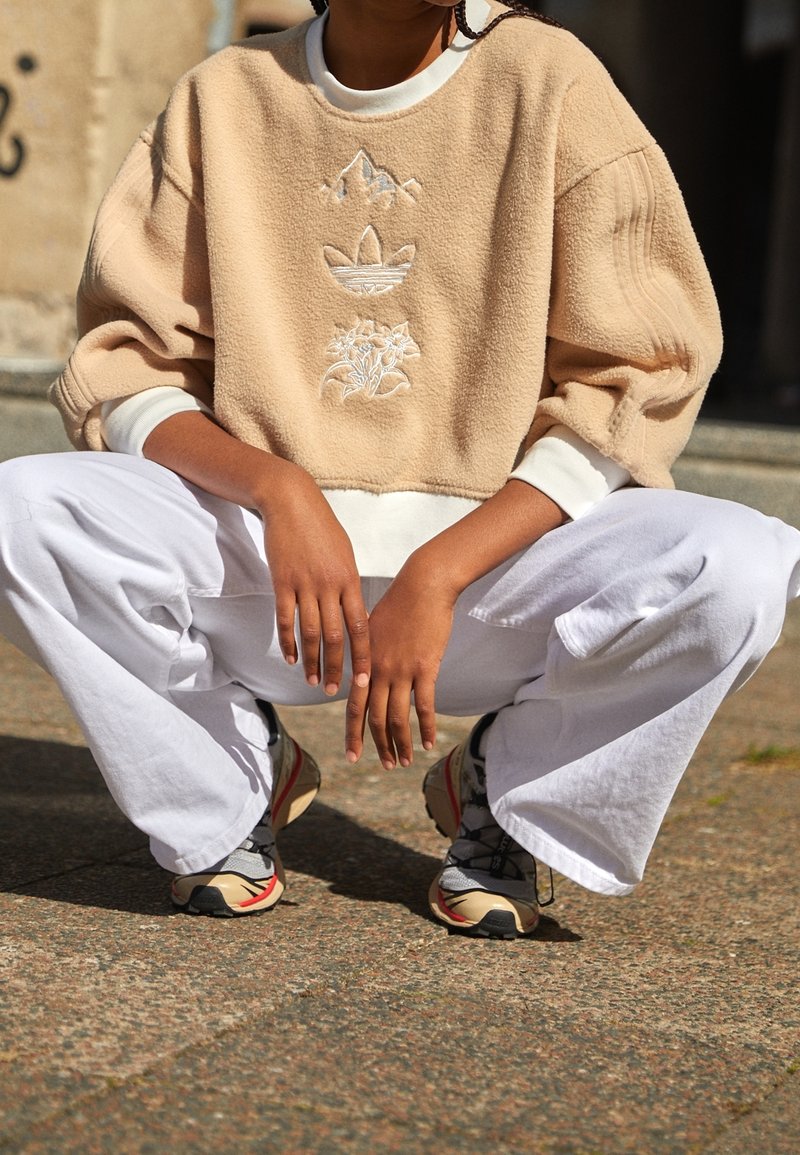 Beige fleece-tröja med vita muddar och broderade bergs- och blommönster, i kombination med vita vida byxor och flerfärgade sneakers.