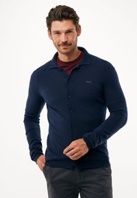Námořnický modrý pletený kardigan s knoflíkovou p�řední částí, dlouhými rukávy a klasickým límcem, který obsahuje malé logo na levé hrudi.