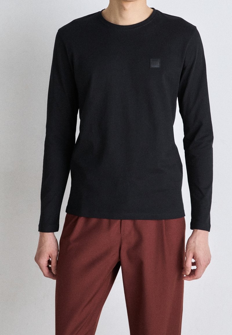 Black long-sleeve t-shirt with a round neckline and a small embossed logo on the chest, paired with dark red trousers.