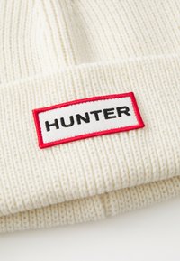 Beanie a coste di colore crema con una toppa rettangolare rossa e bianca che mostra la scritta "HUNTER" in lettere nere sul risvolto.