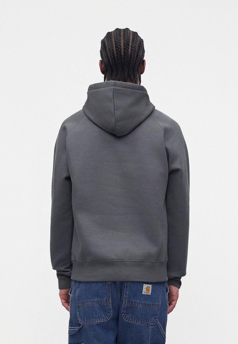 Mann mit geflochtenem Haar, der einen dunkelgrauen Kapuzenpullover und blaue Arbeitshosen trägt, steht mit dem Rücken zu einem schlichten Hintergrund.