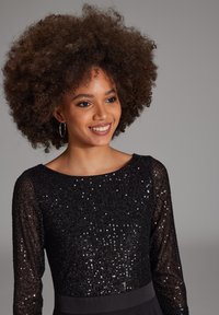Robe noire à sequins avec des manches longues, un col rond et une taille cintrée. Le tissu est brillant avec un aspect texturé dû aux sequins.