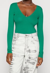 Grüner Langarm-Bodysuit mit tiefem V-Ausschnitt, kombiniert mit ausgebleichten weißen Jeans mit schwarzen Streifen und einem silbernen Knopfverschluss.