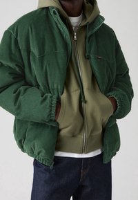 Zielona kurtka sztruksowa z kapturem, pod którą znajduje się oliwkowa bluza z kapturem. Posiada elastyczne mankiety i zamek błyskawiczny, tworząc warstwowy wygląd.