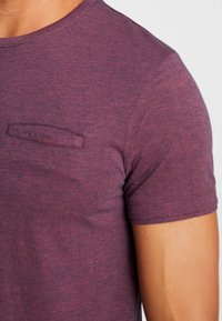 Camiseta de manga corta y cuello redondo en tela sólida de color púrpura oscuro con una textura sutil y un pequeño detalle de bolsillo rectangular en el pecho.