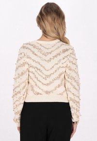 Suéter beige con patrón en zigzag dorado, adornado con cuentas doradas en las mangas, dobladillo acanalado y escote redondo. Textura de punto suave.