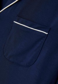 Námořnická modrá tkanina s jemnou texturou, obsahující náprsní kapsu orámovanou bílým lemem. Design je jednoduchý a funkční.