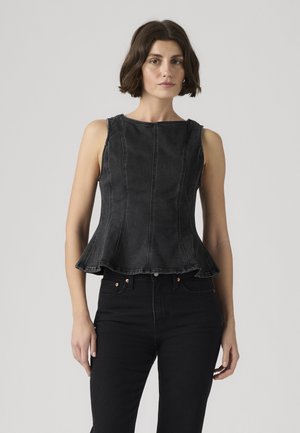 Γυναίκα με κοντά καστανά μαλλιά, φορώντας αμάνικο μαύρο τζιν τοπ με πεπλούμ και μαύρο παντελόνι, στέκεται μπροστά σε απλό ανοιχτόχρωμο φόντο.