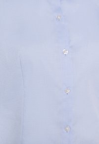 Chemise bleu clair boutonnée en tissu texturé, avec une patte de boutonnage frontale à cinq boutons ronds transparents et un motif à carreaux subtil.