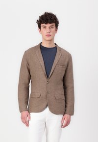 Brun linneblazer med notch-lapel, två framfickor och lätt tygstruktur, bärs över en marinblå skjorta och tillsammans med vita byxor.