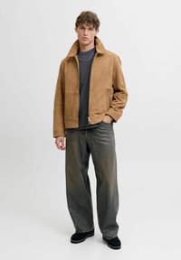 Brun ruskindsjakke med høj krave, lynlåslukning foran og sidelommer; parret med grå løstsiddende jeans og sort fodtøj.