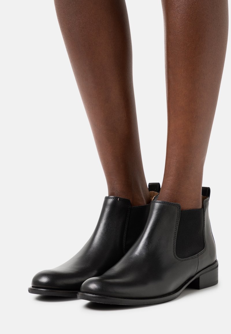 Pieds portant des bottines Chelsea en cuir noir lisse, avec des panneaux latéraux élastiques et des talons blocs bas, sur un fond clair uni.