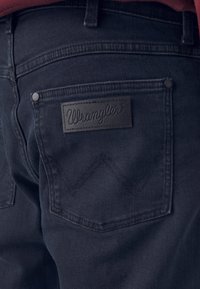 Jeans din denim închis cu un finisaj texturat, având un patch cu logo-ul Wrangler din piele pe buzunarul din spate și un model de cusătură în zigzag.