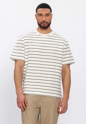 Man met kort haar en baard, gekleed in een wit T-shirt met dunne horizontale olijfgroene strepen en beige broek, tegen een witte achtergrond.