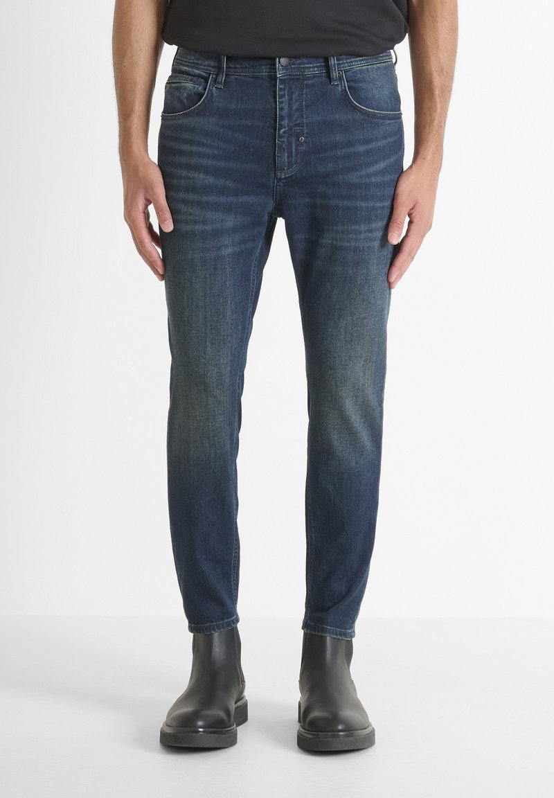 Hombre vestido con jeans ajustados de color azul oscuro y botas de cuero negras hasta el tobillo, de pie contra un fondo blanco liso.