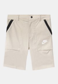 Béžové kraťasy Nike s černými akcenty, které mají dvě boční kapsy a hladkou texturu. Logo je vytištěno v bílé barvě na levém stehně.