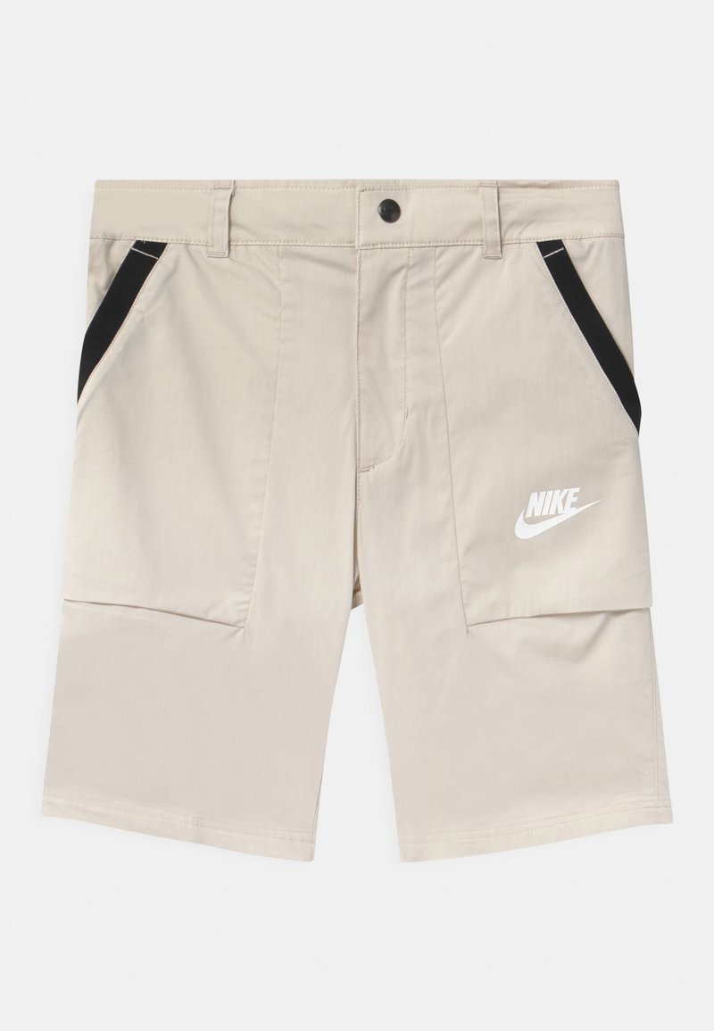 Béžové kraťasy Nike s černými akcenty, které mají dvě boční kapsy a hladkou texturu. Logo je vytištěno v bílé barvě na levém stehně.