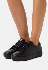 Baskets en cuir noir avec une semelle noire épaisse, dotées d'un bout arrondi et de lacets plats. Marquage visible sur le côté de la semelle.