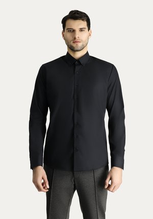 Schwarzes Langarmshirt mit Kragen, Knopfverschluss und glattem Material, kombiniert mit grauen Hosen. Keine Muster oder ausgeprägten Akzente.