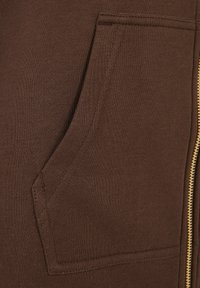 Smeđa zip-up majica s kapuljačom koja se ističe glatkim, teksturiranim materijalom, s prednjim džepom i zlatnim detaljima na zipu.