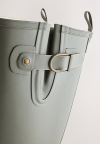 Gros plan d'une botte de pluie en caoutchouc gris avec une sangle réglable à boucle métallique et des languettes de traction sur un fond clair.