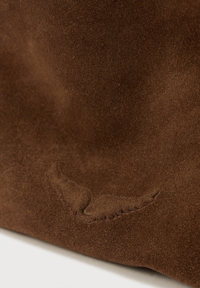 Κοντινό πλάνο καφέ σουέντ υλικού με ανάγλυφη λεπτομέρεια λογότυπου σε σχήμα φτερού κοντά στο κάτω άκρο.