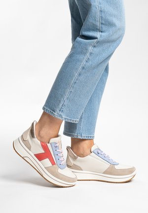 Person in hellblauer Jeans und weißen Turnschuhen mit roten und blauen Akzenten, stehend auf einer weißen Oberfläche.