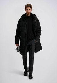 Schwarze Puffy-Jacke mit Kapuze, über einem schwarzen Pullover getragen. Er trägt graue Handschuhe, dunkle karierte Hosen und schwarze Schuhe.