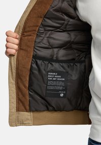 Hånd, der holder en åben beige jakke, der viser en quiltet sort inderside med et sort mærke med hvid tekst indeni en lomme.