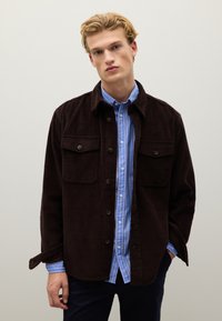 Bruine corduroy jas met twee borstzakken, zwarte knopen en een blauw-wit gestreept shirt eronder. Eenvoudig, casual ontwerp.