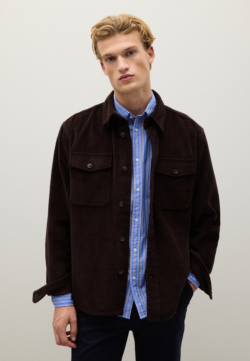 Bruine corduroy jas met twee borstzakken, zwarte knopen en een blauw-wit gestreept shirt eronder. Eenvoudig, casual ontwerp.