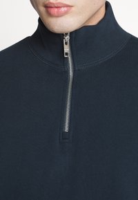 Marinekleurige ritspullover van ribstof. Heeft een hoge kraag en een zilveren rits voor sluiting. Schoon en eenvoudig ontwerp met een gestructureerde afwerking.