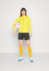 Yellow athletic zip-up jacket, black shorts, orange socks with black stripes, light blue soccer cleats, holding a multicolored soccer ball.