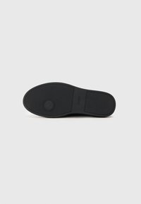 Semelle de chaussure en caoutchouc noir avec un motif de grip texturé et un détail de logo circulaire. Forme arrondie avec une finition lisse.