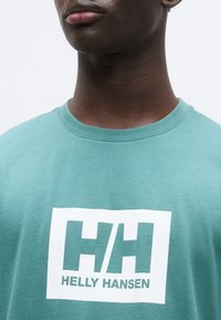 Πράσινη μέντα βαμβακερή κοντομάνικη μπλούζα με ένα λευκό ορθογώνιο λογότυπο που αναγράφει "HH" και "HELLY HANSEN". Στρογγυλή λαιμόκοψη και κοντά μανίκια.