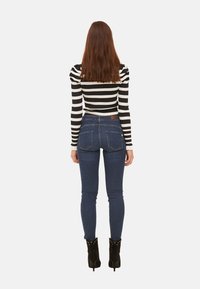 Žena s dlouhými hnědými vlasy, oblečená v černo-bíle pruhovaném topu, tmavě modrých skinny džínách a černých kožených kotníkových botách, stojí zády.