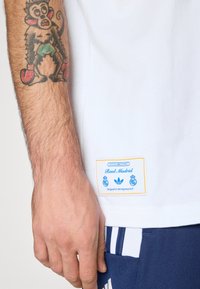 Hvit bomull T-skjorte med en blå og gul etikett med "Real Madrid," og "Designet i Herzogenaurach," på nedre venstre side.