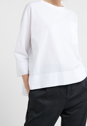 Wit overhemd met lange mouwen en een voorzak, ongestopt gedragen over zwarte gestructureerde broek, bij een persoon die met de handen in de zakken staat.