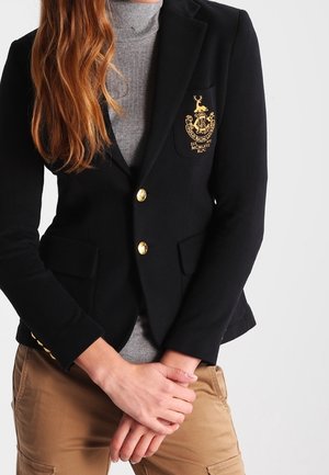 Kobieta ubrana w czarną marynarkę z złotymi guzikami i herbem, pod którą ma szary golf, w połączeniu z beżowymi spodniami typu cargo, trzymająca ręce złączone przed sobą.