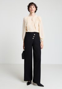 Blusa crema con dettagli in pizzo arricciato e collo annodato; pantaloni neri a vita alta con bottoni laterali; eleganti scarpe nere con punta affusolata.