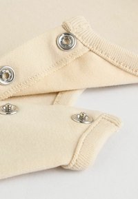 Helle beige Stoff mit metallischen Druckknöpfen und gerippten Bündchen. Glatte Textur und einfaches Design mit sichtbaren Nähdetails.