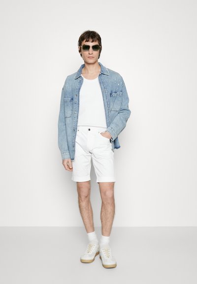 Lys blå denimjakke, hvit tank topp, hvite shorts, hvite joggesko med beige såle og mørke solbriller; satt mot en ensfarget bakgrunn.