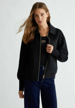 Giacca nera con zip, presenta un design con colletto, tasche frontali e polsini a costine. Abbinata a pantaloni in denim scuro.