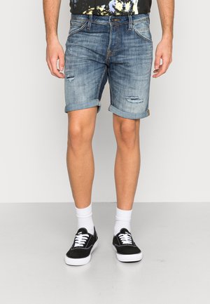 Man iförda upprullade blå denimshorts med slitna lappar, vita ankelsockor och svarta låga sneakers stående på ett grått golv.