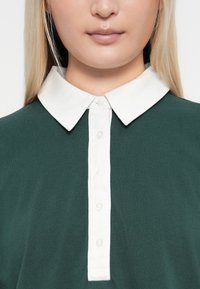Grön skjorta med krage och vit knappslå. Mjuk, texturerad tyg. Fem knappar synliga längs knappslån.